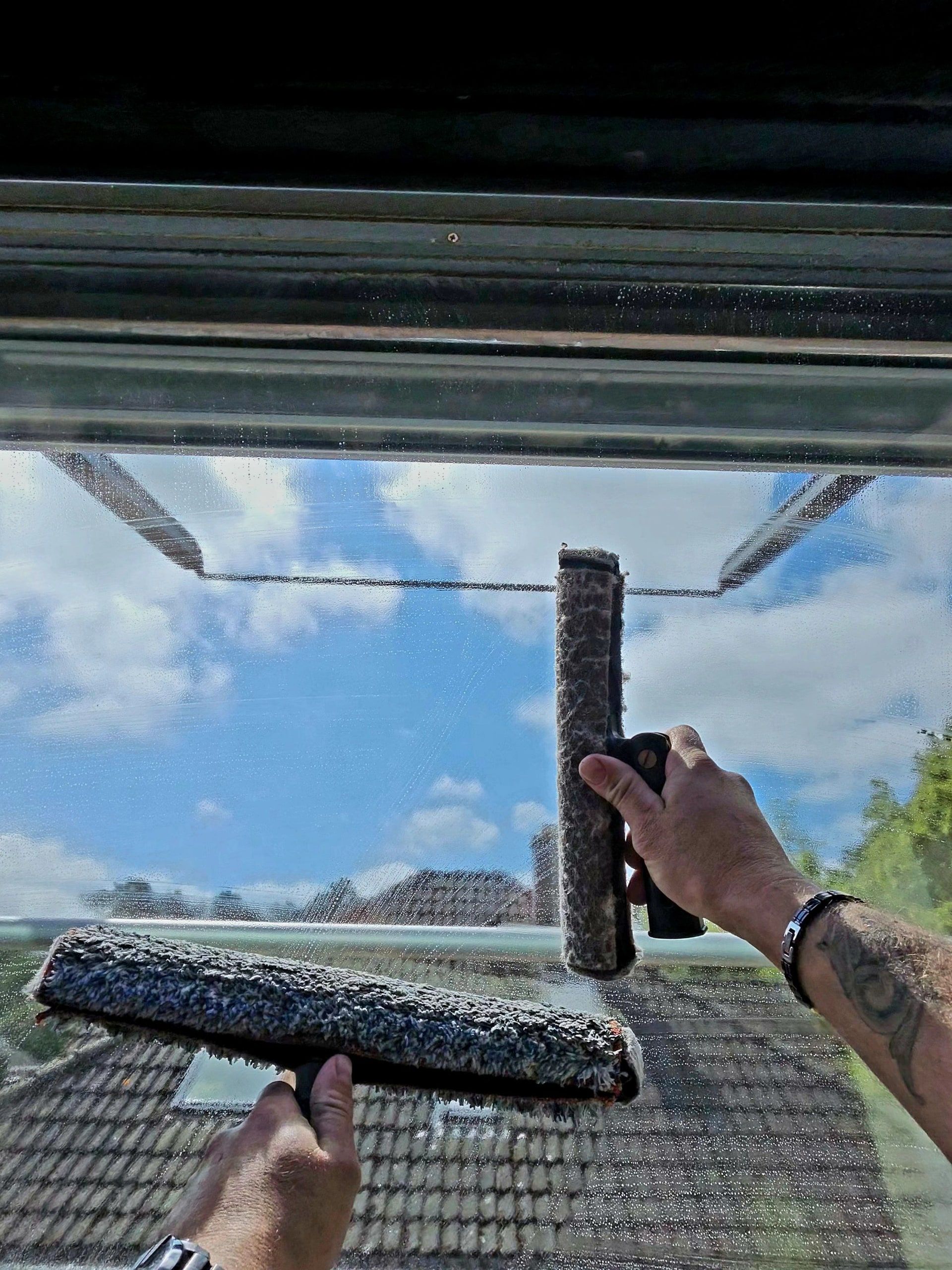 Zwei Einwascher verteilen Reinigungswasser auf einer großen Fensterscheibe, in der sich blauer Himmel mit Wolken spiegelt