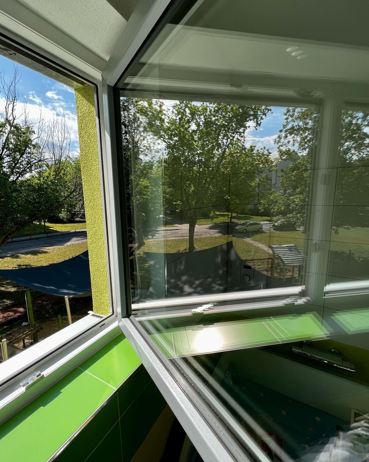Geöffnetes Eckfenster mit Blick auf Bäume und einen Spielplatz, davor bunte Fensterbänke