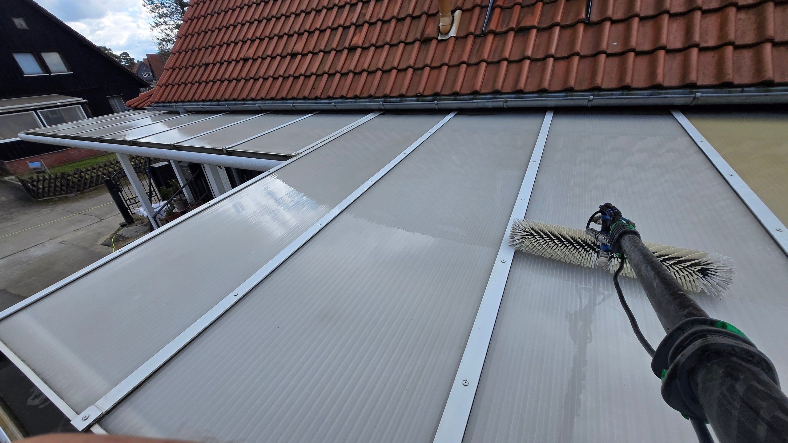 Reinwasserbürste an der Carbonstange reinigt die hellen Stegplatten einer Terrassenüberdachung; oben schließt das rote Ziegeldach des Hauses an, in den bereits gereinigten Feldern spiegelt sich der Himmel