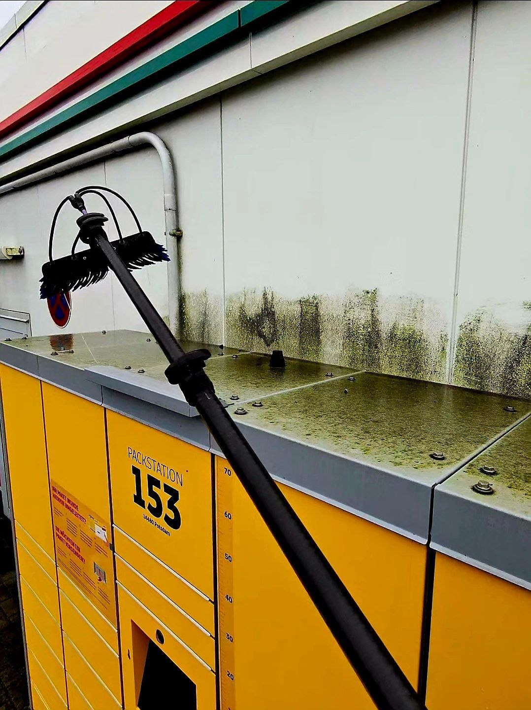 Reinigungsbürste entfernt Algenbewuchs von der Metallabdeckung einer DHL-Packstation
