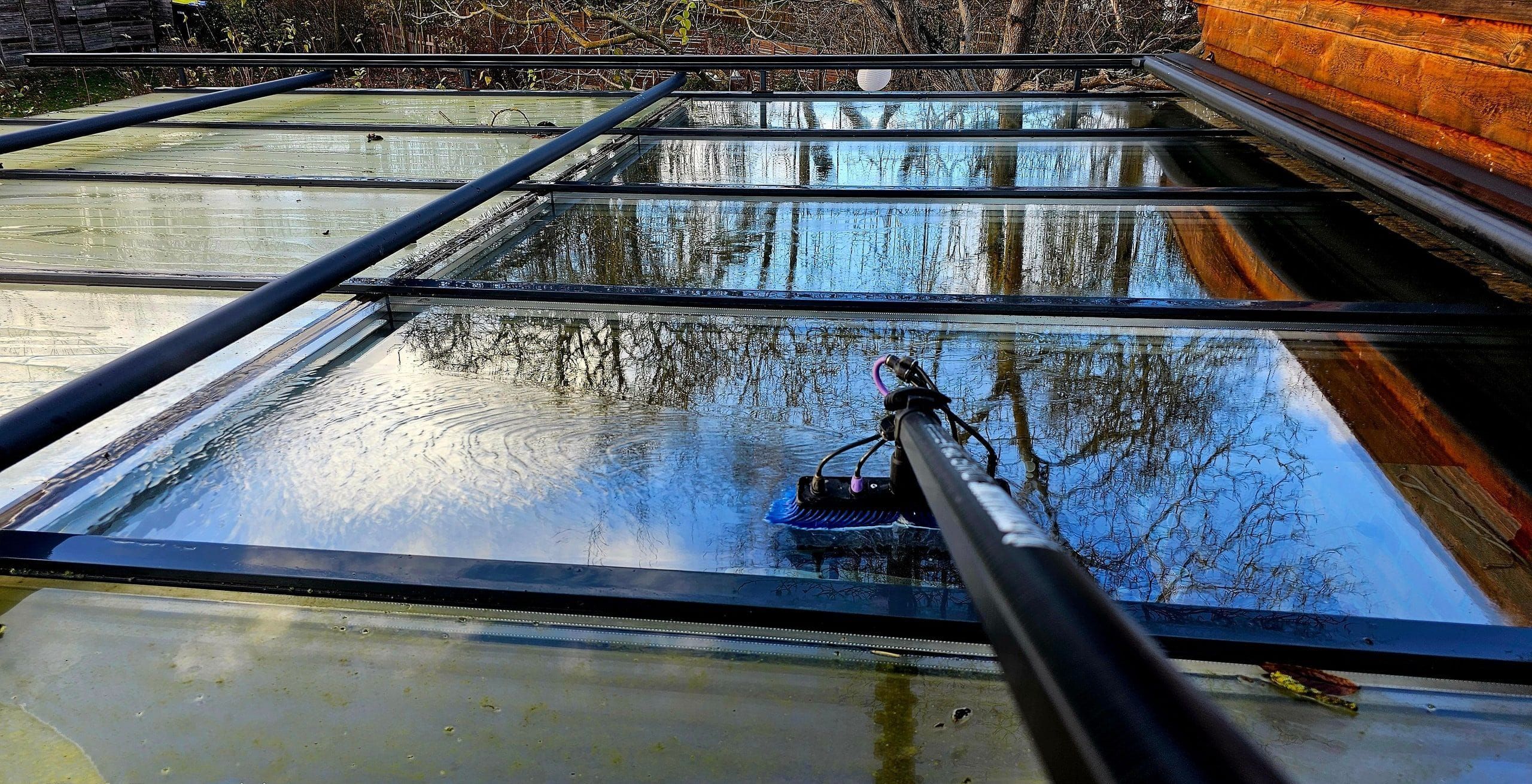 Nasses Glasdach einer Terrassenüberdachung während der Reinigung: die blaue Reinwasserbürste an der Carbonstange liegt auf der Scheibe, im ablaufenden Wasserfilm spiegeln sich kahle Bäume und blauer Himmel