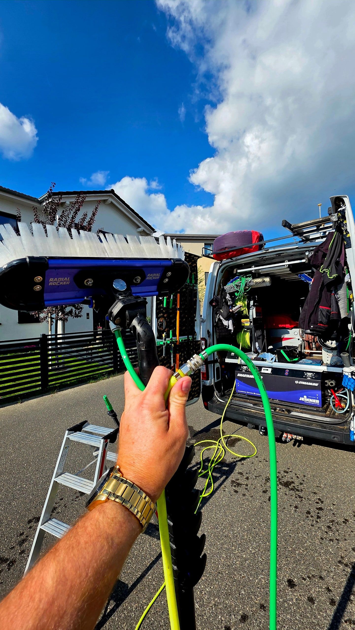 Grüner Wasserschlauch mit Messingkupplung in der Hand, dahinter der Bürstenkopf an der Stange, eine Leiter und das geöffnete Einsatzfahrzeug