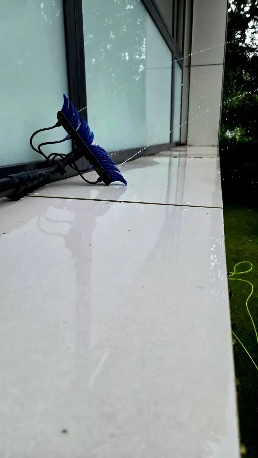 Dieselbe Fensterbank nach der Reinigung vom selben Standpunkt: der Beton ist hell, belagfrei und nass glänzend, an der Scheibe steht die blaue Reinwasserbürste an der Carbonstange und sprüht entmineralisiertes Wasser