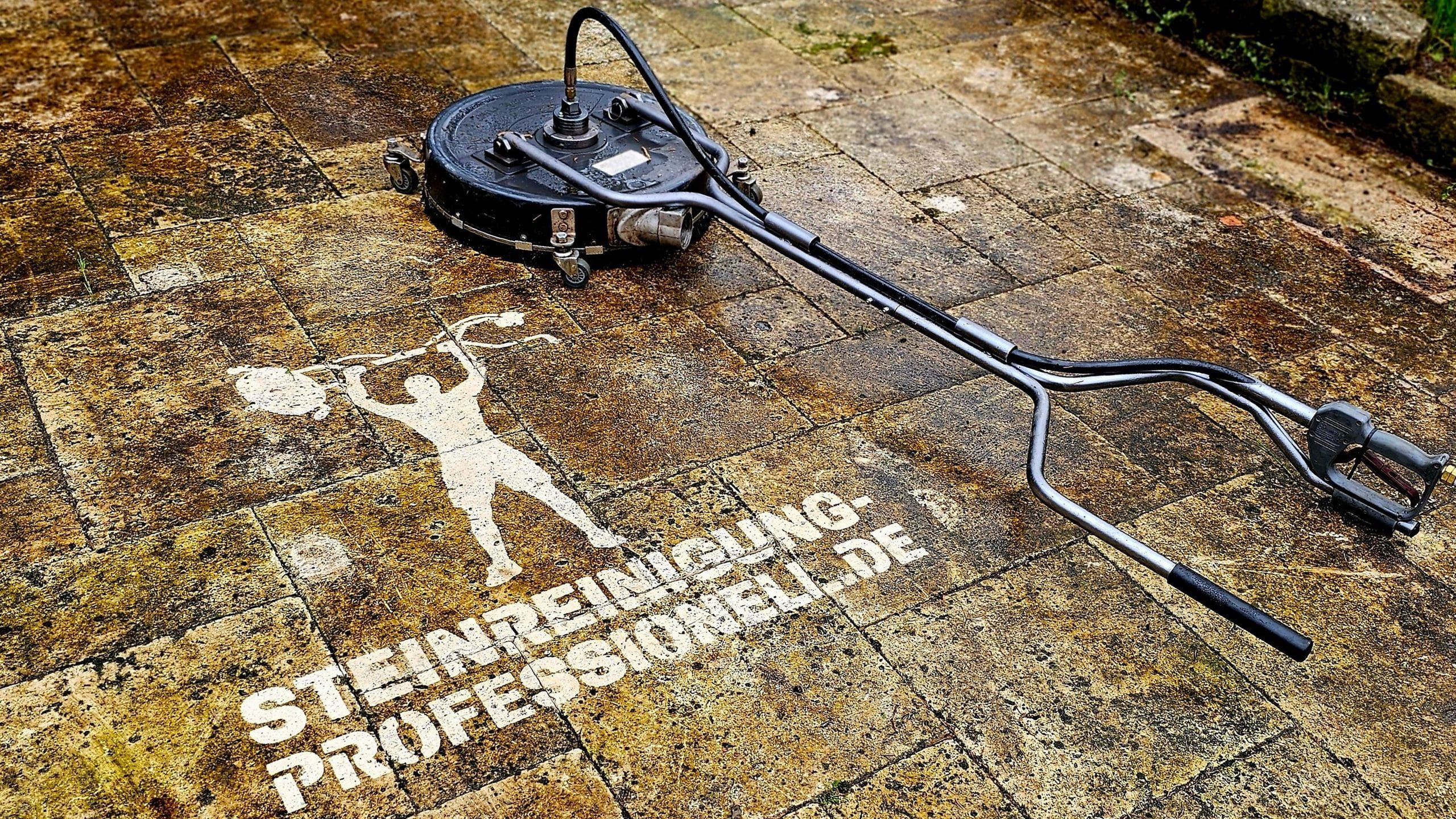 Runder Flächenreiniger mit Carbonlanze auf einer stark veralgten Natursteinfläche; daneben ist der Schriftzug „Steinreinigung-Professionell.de" samt Figur eines Gewichthebers sauber in den grün-braunen Belag hineingereinigt, sodass die helle Steinfarbe darunter die Buchstaben bildet