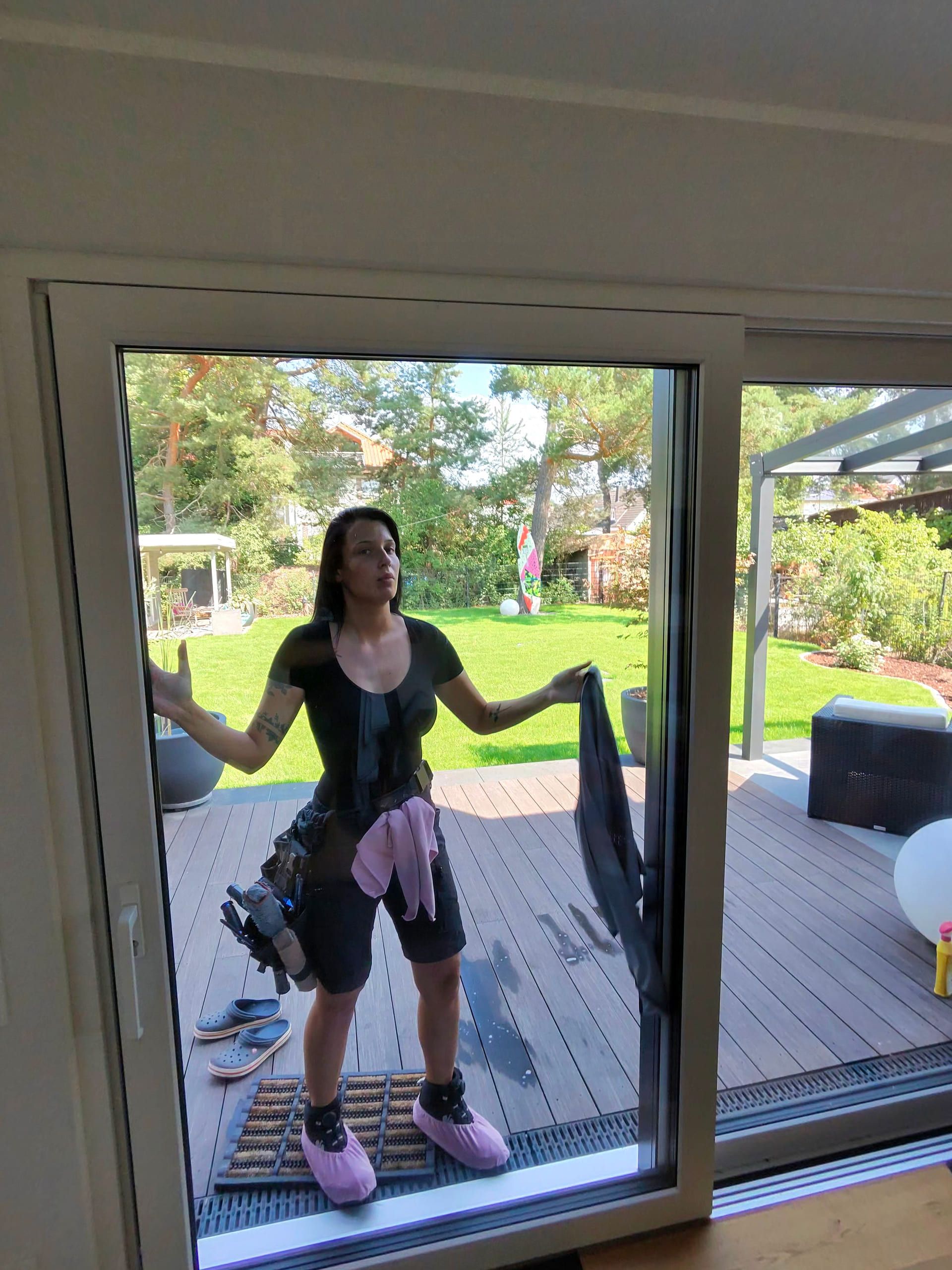 Annabell reinigt von der Terrasse aus die Scheibe einer bodentiefen Glasschiebetür; die Aufnahme entsteht von innen durch das Glas
